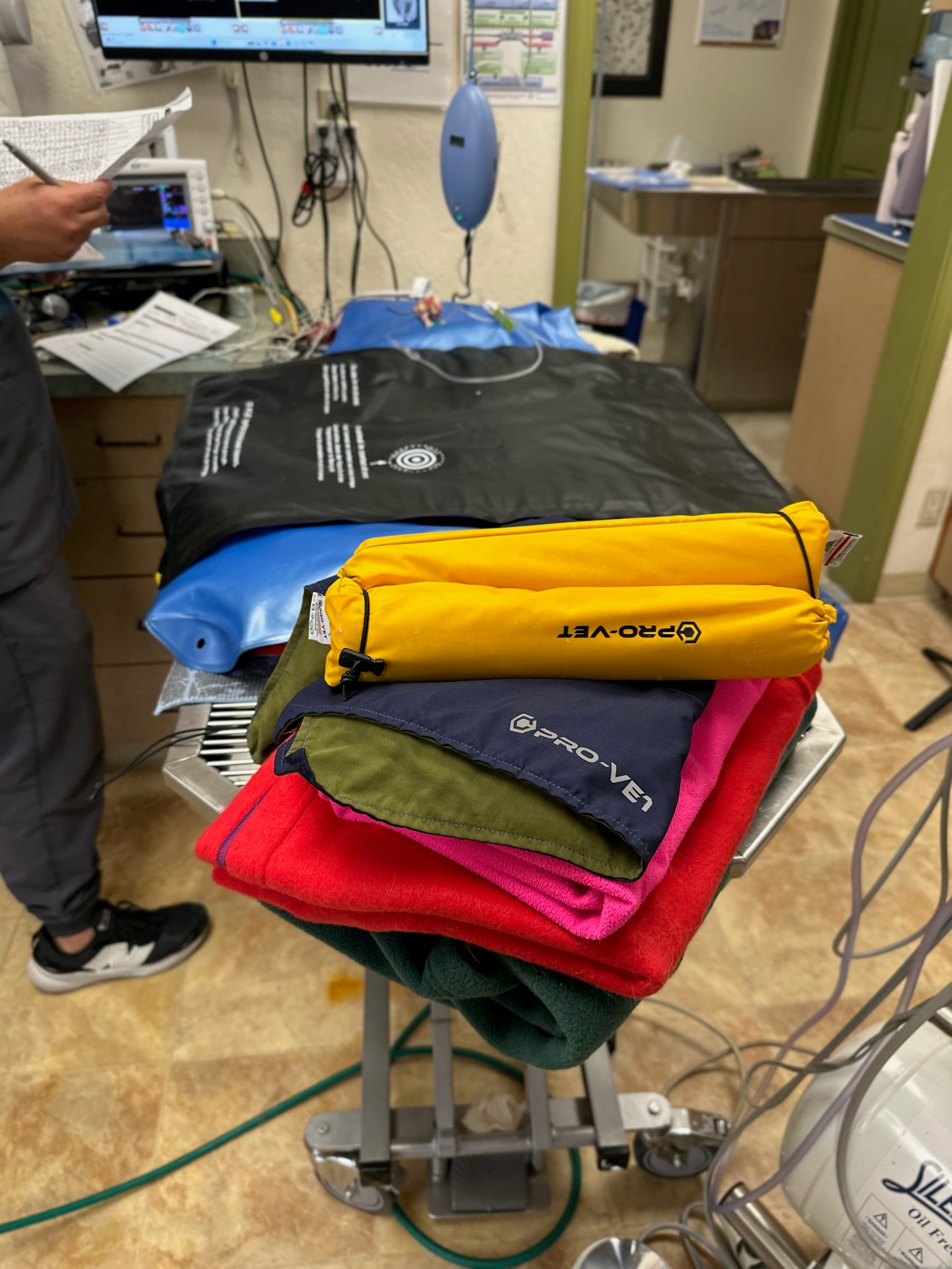 A stack of folded Pro-Vet cold therapy packs in various colors including yellow, navy, green, red, and blue, placed on a metal treatment table inside what appears to be an office or clinic room.